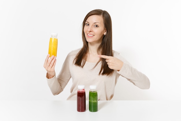 Jovem mulher à mesa com smoothies de desintoxicação verdes, vermelhos e amarelos em garrafas isoladas no fundo branco. Nutrição adequada, bebida vegetariana, estilo de vida saudável, conceito de dieta. Área com espaço de cópia.