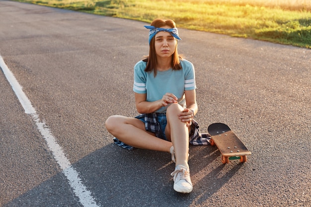 Jovem mulher adulta morena sentada na estrada de asfalto tendo trauma após cair do skate, tendo dor no joelho, olhando para a câmera com o rosto carrancudo.