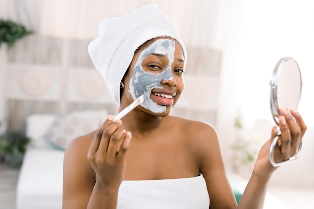 Jovem mulher africana bonita toalha branca, aplicação de máscara facial e segurando o espelho. Conceito de cuidados com a pele Spa