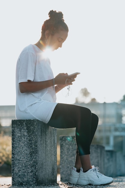 Jovem mulher africana branca grande sede verificando o telefone depois de um dia de exercícios sentado perto da estrada durante o pôr do sol. Mulher cansada de fitness suando, fazendo uma pausa para ouvir música no telefone.