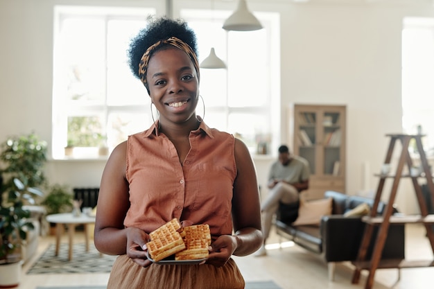 Jovem mulher africana feliz com waffles caseiros olhando para você com um sorriso cheio de dentes ao lado do marido sentado no sofá