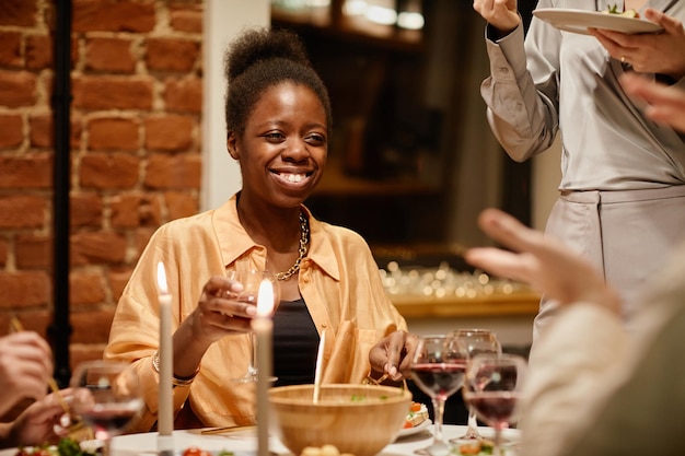 Jovem mulher afro-americana alegre com copo de vinho tinto curtindo a festa em casa enquanto está sentado perto de si