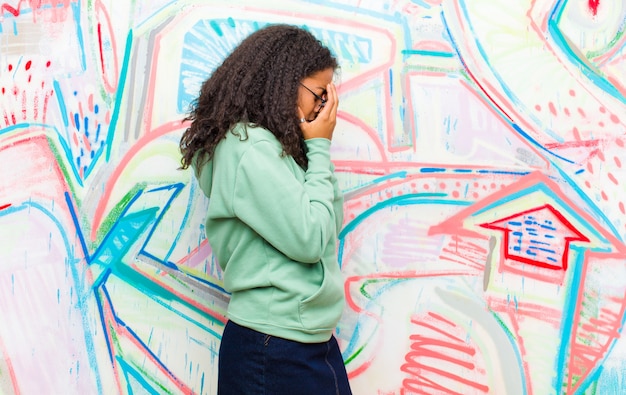 Jovem mulher afro-americana bonita cobrindo os olhos com as mãos com um olhar triste e frustrado de desespero, chorando contra a parede do graffiti