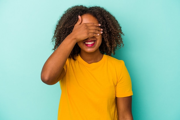 Jovem mulher afro-americana com cabelo encaracolado isolado em um fundo azul cobre os olhos com as mãos, sorri amplamente esperando por uma surpresa.