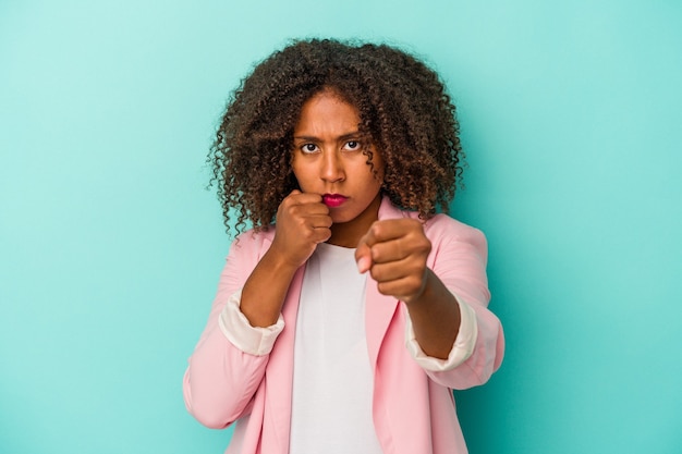 Jovem mulher afro-americana com cabelo encaracolado, isolado em um fundo azul, dando um soco, raiva, lutando devido a uma discussão, boxe.