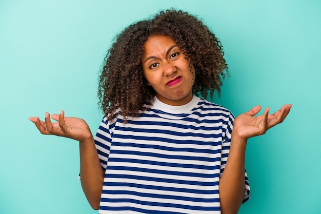 Jovem mulher afro-americana com cabelo encaracolado, isolado em um fundo azul, duvidando e encolhendo os ombros em gesto de questionamento.