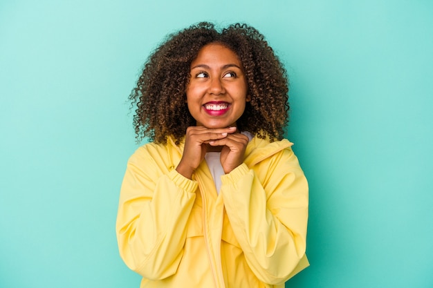 Jovem mulher afro-americana com cabelo encaracolado, isolado em um fundo azul mantém as mãos sob o queixo, olhando alegremente para o lado.