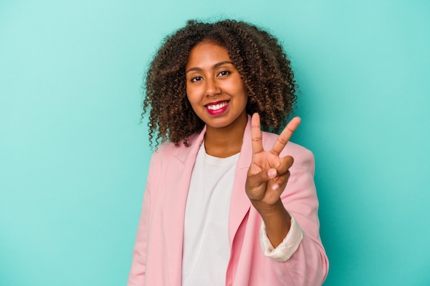 Jovem mulher afro-americana com cabelo encaracolado, isolado em um fundo azul, mostrando o sinal da vitória e sorrindo amplamente.