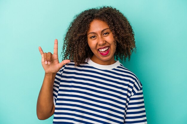 Jovem mulher afro-americana com cabelo encaracolado, isolado em um fundo azul, mostrando um gesto de chifres como um conceito de revolução.