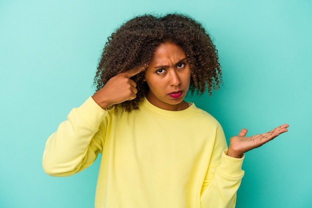 Jovem mulher afro-americana com cabelo encaracolado, isolado em um fundo azul, mostrando um gesto de decepção com o dedo indicador.