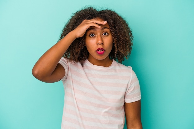 Jovem mulher afro-americana com cabelo encaracolado, isolado em um fundo azul, olhando para longe, mantendo a mão na testa.