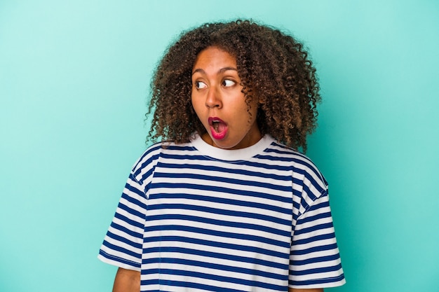 Jovem mulher afro-americana com cabelo encaracolado, isolado em um fundo azul, sendo chocada por causa de algo que ela viu.