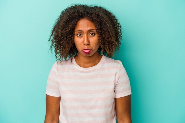 Jovem mulher afro-americana com cabelo encaracolado, isolado no fundo azul, rosto triste, sério, sentindo-se miserável e descontente.