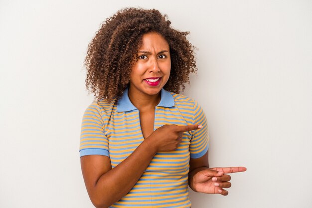 Jovem mulher afro-americana com cabelo encaracolado, isolado no fundo branco chocada, apontando com o dedo indicador para um espaço de cópia.