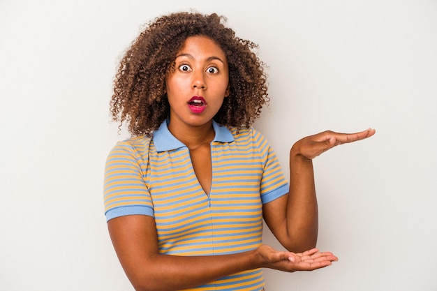 Jovem mulher afro-americana com cabelo encaracolado, isolado no fundo branco chocada e espantada, segurando um espaço de cópia entre as mãos.