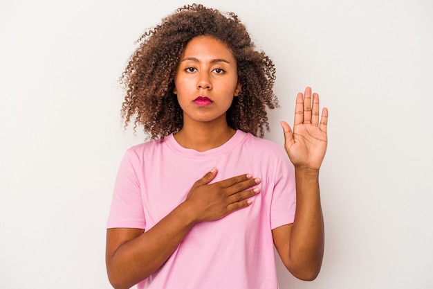 Foto jovem mulher afro-americana com cabelo encaracolado, isolado no fundo branco, fazendo um juramento, colocando a mão no peito.