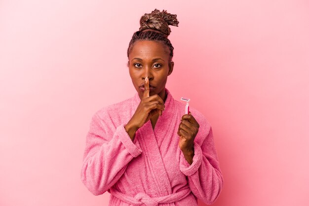 Jovem mulher afro-americana com roupão rosa segurando a lâmina de barbear isolada no fundo rosa, mantendo um segredo ou pedindo silêncio.