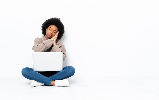 Jovem mulher afro-americana com um laptop sentado no chão, fazendo o gesto do sono em expressão dorable