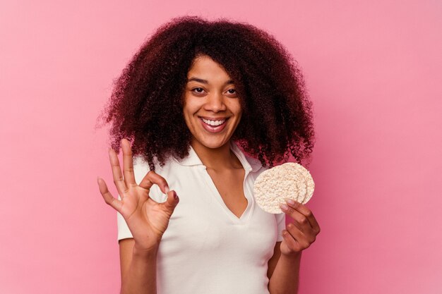 Jovem mulher afro-americana comendo um bolo de arroz isolado na parede rosa alegre e confiante, mostrando o gesto de ok.