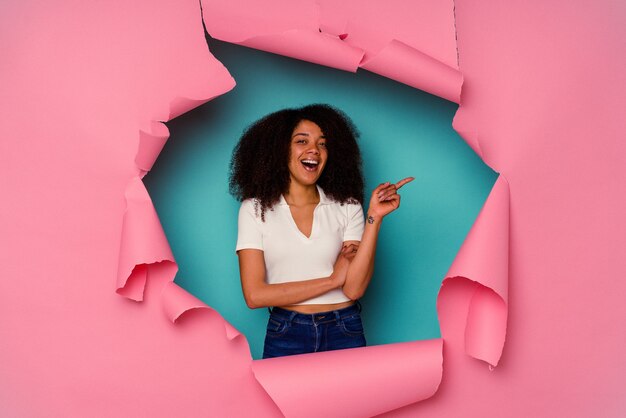 Jovem mulher afro-americana em papel rasgado, isolado em azul, sorrindo alegremente, apontando com o dedo indicador para longe.