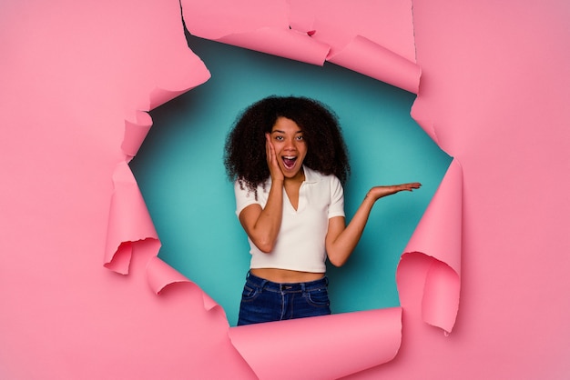 Jovem mulher afro-americana em papel rasgado, isolado em um fundo azul, tem espaço de cópia na palma da mão, mantenha a mão na bochecha Espantado e encantado.