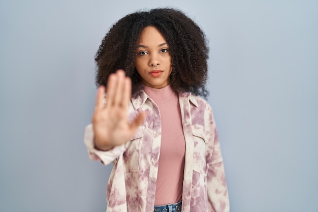 Jovem mulher afro-americana em pé sobre fundo azul fazendo parar de cantar com a palma da mão. expressão de advertência com gesto negativo e sério no rosto.