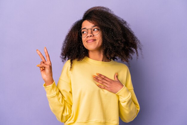 Jovem mulher afro-americana encaracolada isolada na parede roxa, fazendo um juramento, colocando a mão no peito.