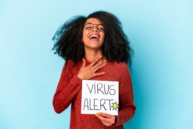 Jovem mulher afro-americana encaracolada segurando um cartaz de alerta de vírus ri alto, mantendo a mão no peito.