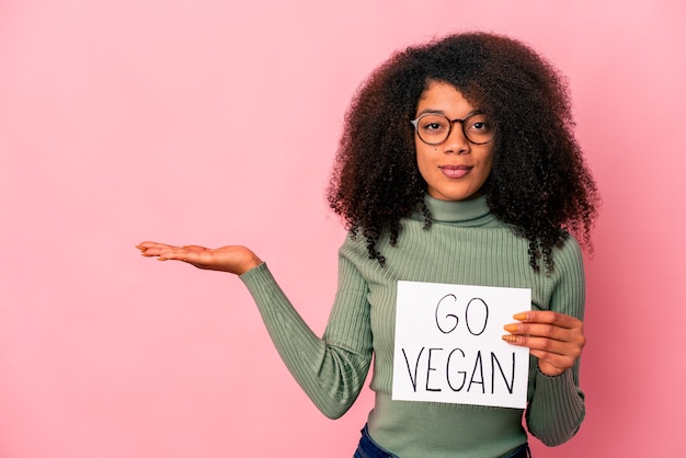 Jovem mulher afro-americana encaracolada segurando um cartaz de veganos mostrando um espaço de cópia na palma da mão e segurando a outra mão na cintura.