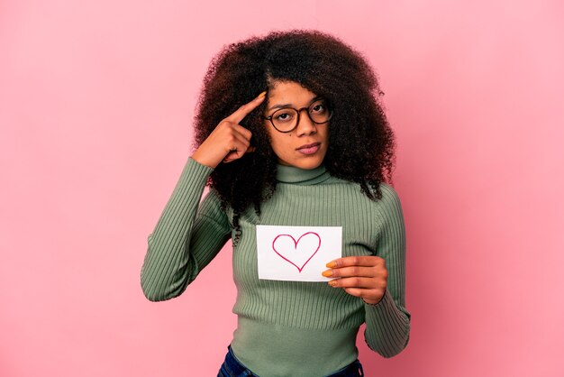 Jovem mulher afro-americana encaracolada segurando um símbolo do coração no cartaz apontando o templo com o dedo, pensando, focado em uma tarefa.