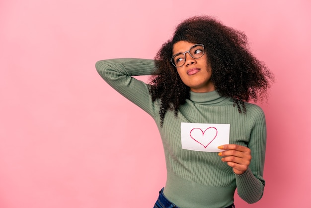 Jovem mulher afro-americana encaracolada segurando um símbolo do coração no cartaz tocando a parte de trás da cabeça, pensando e fazendo uma escolha.