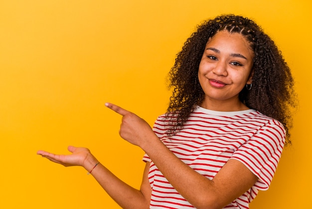 Jovem mulher afro-americana isolada em fundo amarelo animado segurando um espaço de cópia na palma da mão.