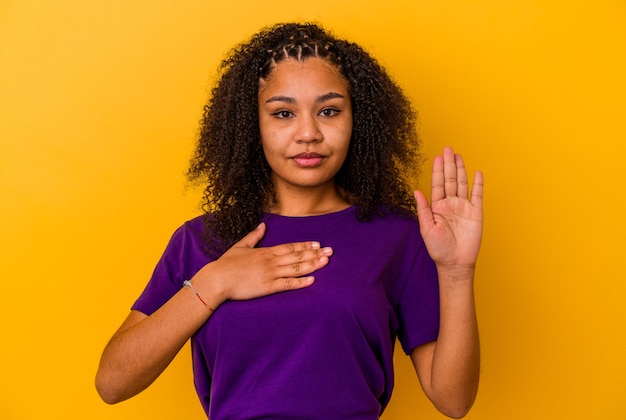 Jovem mulher afro-americana isolada em fundo amarelo, fazendo um juramento, colocando a mão no peito.