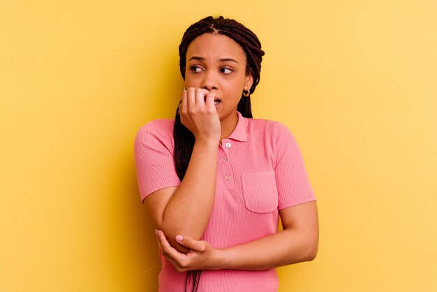 Jovem mulher afro-americana isolada em fundo amarelo, roendo as unhas, nervosa e muito ansiosa.