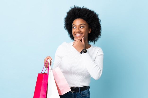 Jovem mulher afro-americana isolada em fundo azul segurando sacolas de compras e pensando