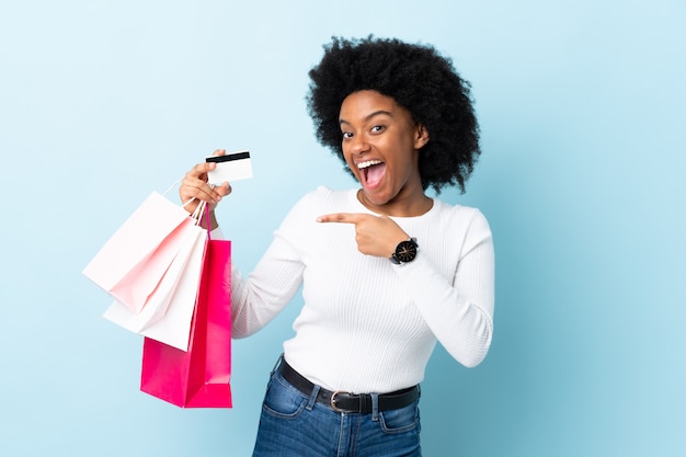 Jovem mulher afro-americana isolada em fundo azul segurando sacolas de compras e um cartão de crédito