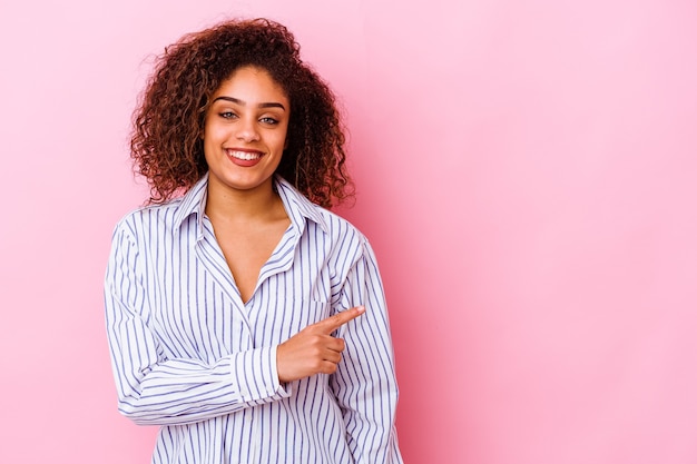 Jovem mulher afro-americana isolada em rosa sorrindo e apontando de lado, mostrando algo no espaço em branco.