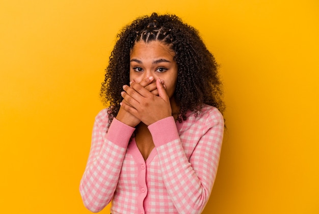 Jovem mulher afro-americana isolada em um fundo amarelo, cobrindo a boca com as mãos parecendo preocupadas.