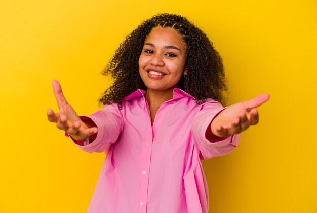 Jovem mulher afro-americana isolada em um fundo amarelo, mostrando uma expressão de boas-vindas.