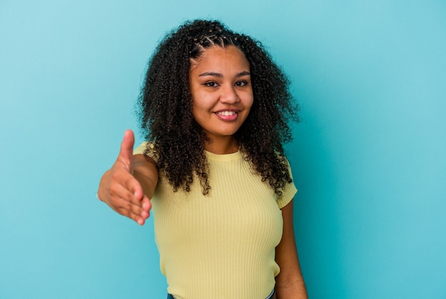 Jovem mulher afro-americana isolada em um fundo azul, esticando a mão na câmera em um gesto de saudação.