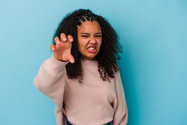 Jovem mulher afro-americana isolada em um fundo azul, mostrando garras imitando um gato, gesto agressivo.