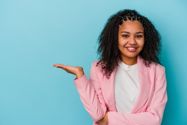 Jovem mulher afro-americana isolada em um fundo azul, mostrando um espaço de cópia na palma da mão e segurando a outra mão na cintura.