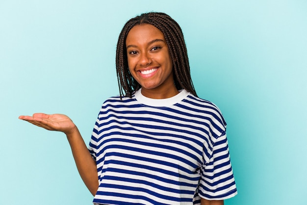 Jovem mulher afro-americana isolada em um fundo azul, mostrando um espaço de cópia na palma da mão e segurando a outra mão na cintura.