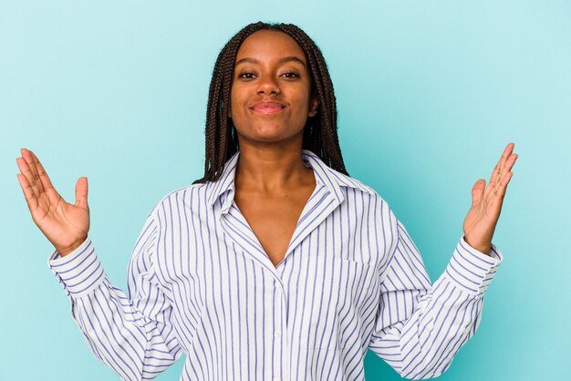 Jovem mulher afro-americana isolada em um fundo azul, segurando algo pequeno com os indicadores, sorrindo e confiante.