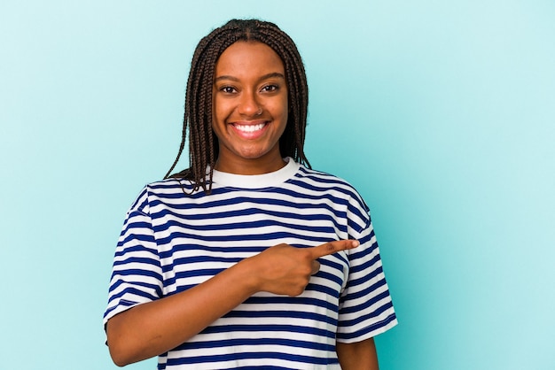 Jovem mulher afro-americana isolada em um fundo azul, sorrindo e apontando de lado, mostrando algo no espaço em branco.