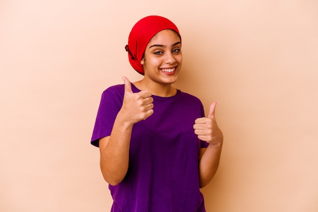 Jovem mulher afro-americana isolada em um fundo bege, levantando os dois polegares, sorrindo e confiante.