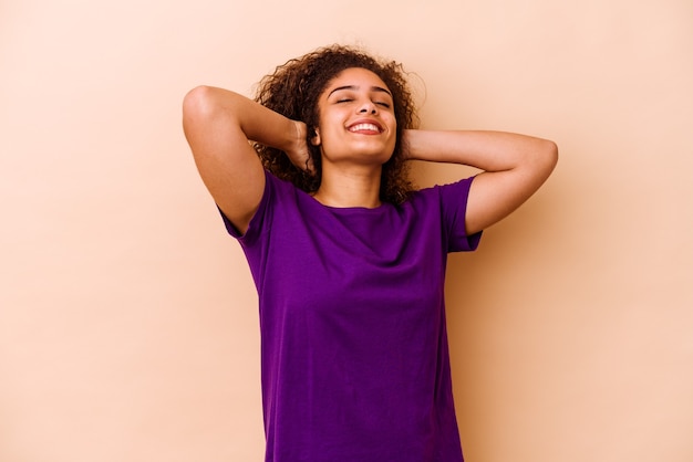 Jovem mulher afro-americana isolada em um fundo bege, sentindo-se confiante, com as mãos atrás da cabeça.