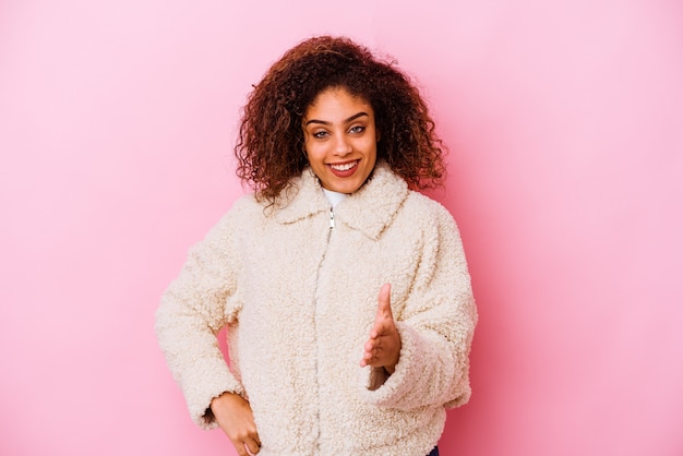 Jovem mulher afro-americana isolada em um fundo rosa, esticando a mão na câmera em um gesto de saudação.