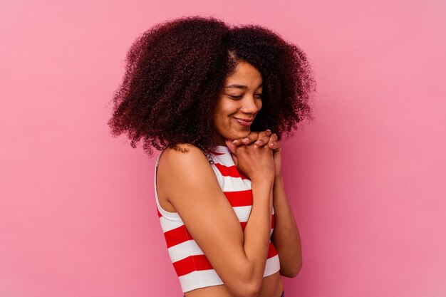 Jovem mulher afro-americana isolada em um fundo rosa mantém as mãos sob o queixo, olhando alegremente para o lado.