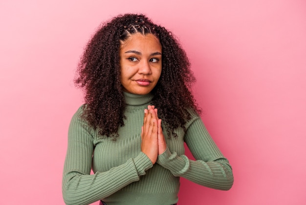 Jovem mulher afro-americana isolada em um fundo rosa orando, mostrando devoção, pessoa religiosa em busca de inspiração divina.